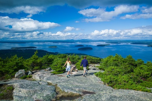A couple walks along a rocky trail overlooking a scenic view of islands and a blue sky with clouds.
