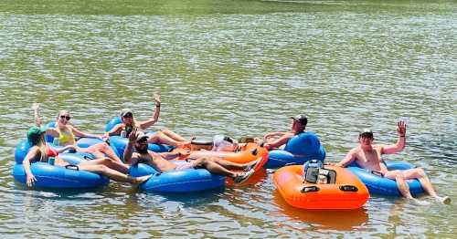 A group of people relaxes on colorful inflatable tubes in a calm river, waving and enjoying a sunny day.