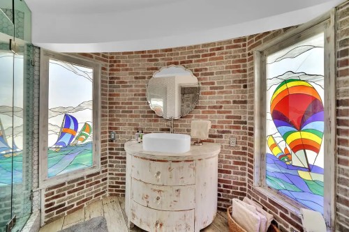 A stylish bathroom with brick walls, a round mirror, and colorful stained glass windows featuring sailboats and hot air balloons.