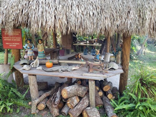 A rustic outdoor kitchen with a thatched roof, featuring wooden tables, crafts, and decorative items.