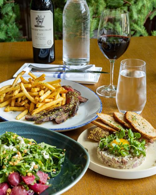 A table setting with a steak, fries, salad, tartare, wine, and water, surrounded by greenery.
