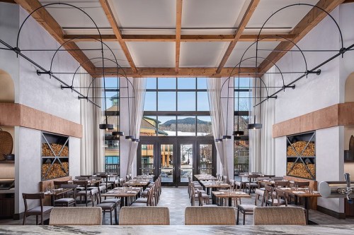 Modern restaurant interior with wooden beams, large windows, and neatly arranged tables. Mountain view in the background.