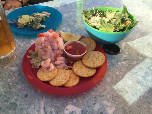 A red plate with shrimp, crackers, and cocktail sauce, alongside a blue bowl of salad and a glass of beer.