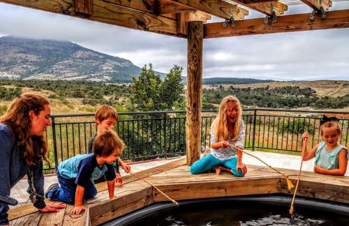 A group of children and an adult are fishing with sticks at a wooden deck overlooking a scenic landscape.