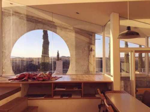 Modern interior with large circular window, wooden surfaces, and a view of trees and landscape outside. Warm lighting.