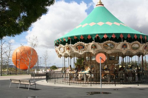 A colorful carousel with horses, set against a backdrop of a large orange sphere and cloudy sky.