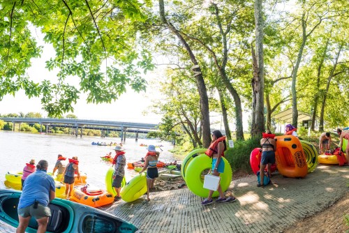 A group of people preparing colorful kayaks and inflatable rafts by a river under sunny trees.