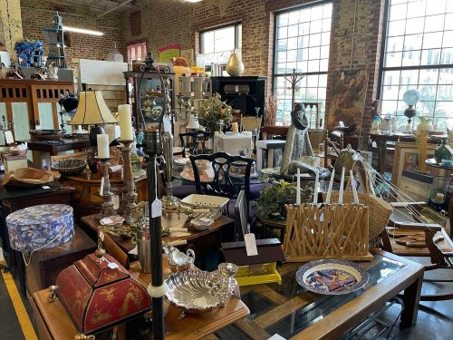 A cluttered antique shop filled with various vintage items, furniture, and decorative pieces. Bright windows in the background.