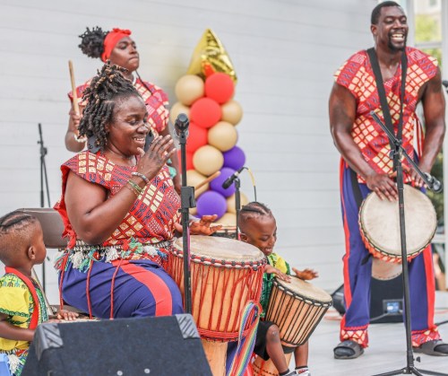 A lively performance featuring musicians in colorful outfits playing drums, with children participating on stage.