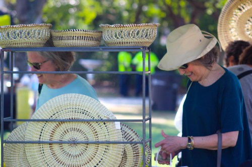 Two women browse woven baskets at an outdoor market, with a display of baskets in the foreground.