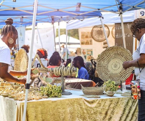 Two women display and sell handmade woven crafts at a market under a blue tent.