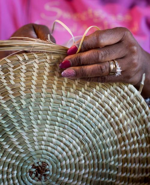 A close-up of hands weaving a circular basket, showcasing intricate patterns and colorful nails.