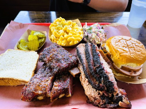 A plate of barbecue ribs, sliced meat sandwich, macaroni, coleslaw, pickles, and bread on a red tray.