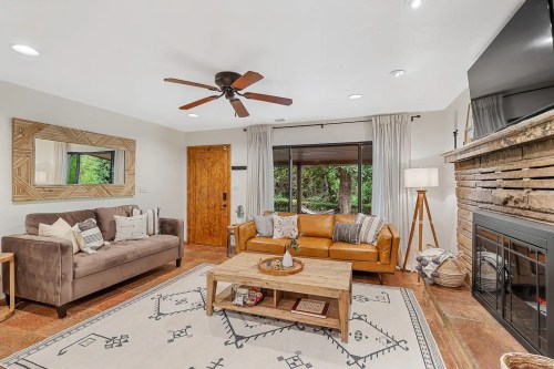 Cozy living room with a brown leather sofa, gray couch, wooden coffee table, and a stone fireplace, surrounded by greenery.