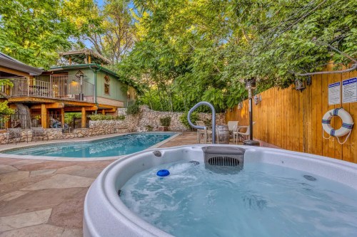 A hot tub beside a swimming pool, surrounded by trees and a wooden fence, with a cozy house in the background.