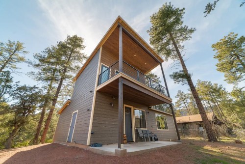 A modern two-story house surrounded by tall trees, featuring a balcony and large windows.