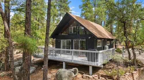 A modern black cabin with a deck, surrounded by tall trees and rocky landscaping in a serene outdoor setting.