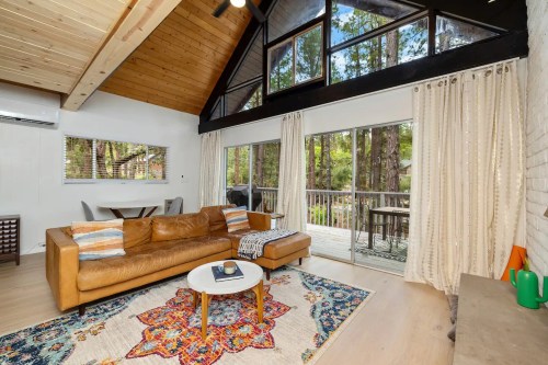 Cozy living room with a brown leather sofa, colorful rug, large windows, and a view of trees outside.