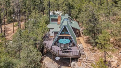 Aerial view of a modern A-frame cabin surrounded by tall trees and a deck with a green umbrella.