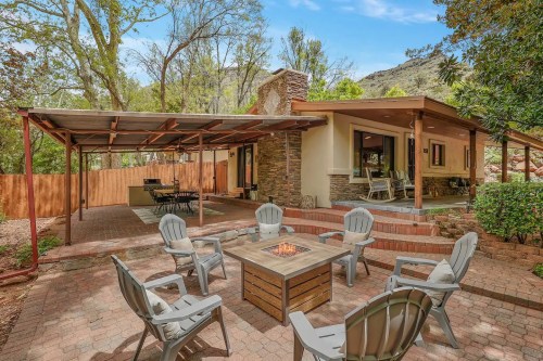A cozy outdoor patio with a fire pit, surrounded by chairs, and a house with a stone chimney in a wooded area.