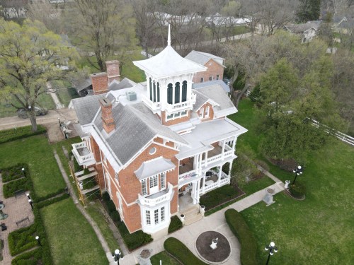 Aerial view of a large, historic brick mansion with a tower, surrounded by green lawns and trees.