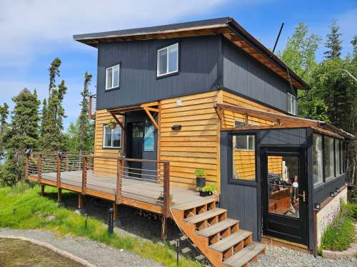 A modern two-story cabin with a wooden exterior, large windows, and a deck surrounded by greenery.
