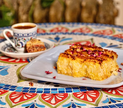 A slice of golden dessert topped with rose petals, next to a cup of coffee on a colorful tiled table.