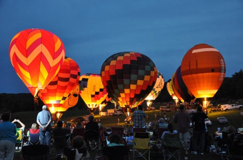 Colorful hot air balloons illuminated at dusk, with spectators seated in front, enjoying the vibrant display.
