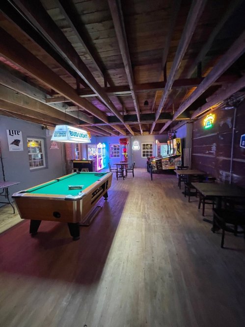 A dimly lit bar interior featuring a pool table, tables, and neon signs on the walls.