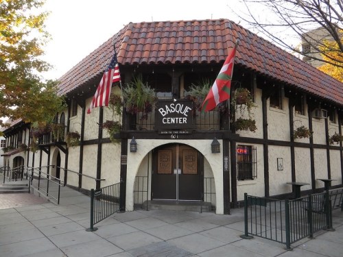 Exterior of the Basque Center, featuring a tiled roof, flags, and decorative plants, in a charming architectural style.
