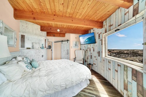 Cozy bedroom with wooden beams, a large window, and a TV, featuring light-colored decor and a beachy vibe.