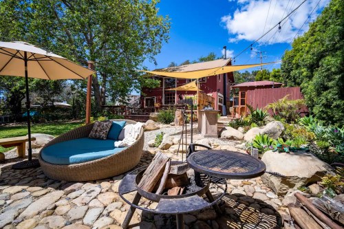 A cozy outdoor space with a circular lounge chair, fire pit, and lush greenery, featuring a red house in the background.