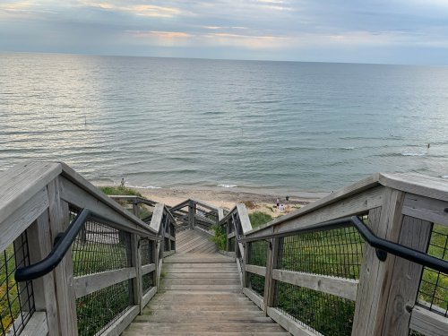 Wooden stairs lead down to a sandy beach by a calm lake, with gentle waves and a cloudy sky in the background.