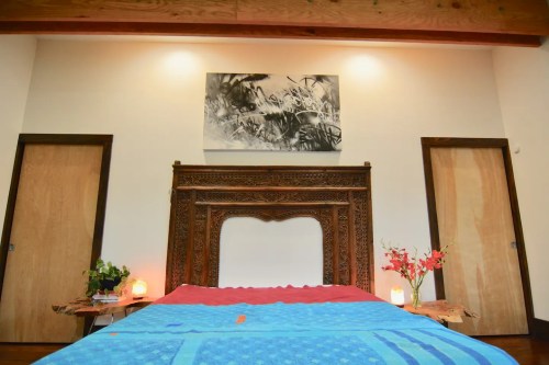 A cozy bedroom featuring a wooden bed frame, colorful bedding, and artwork on the wall, with plants and soft lighting.