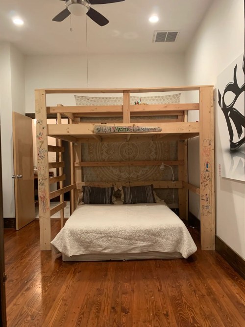 A wooden loft bed with a cozy white bedding set, featuring a decorated wall and wooden floors in a bright room.