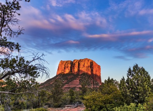 A red rock formation rises against a blue sky with clouds, surrounded by green trees and shrubs.