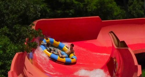 A child rides a colorful inflatable tube down a steep water slide, splashing water around. Lush greenery surrounds the slide.