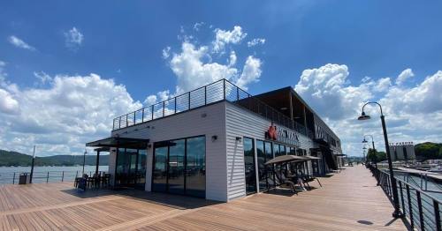A modern waterfront building with large windows and a wooden deck, under a bright blue sky with fluffy clouds.