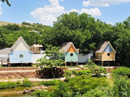 Colorful, whimsical houses with circular windows, set among greenery and a small water feature under a blue sky.