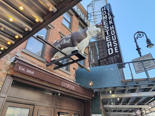 Sign for "Old Homestead Steakhouse" featuring a cow statue, with a vintage marquee and brick building in the background.