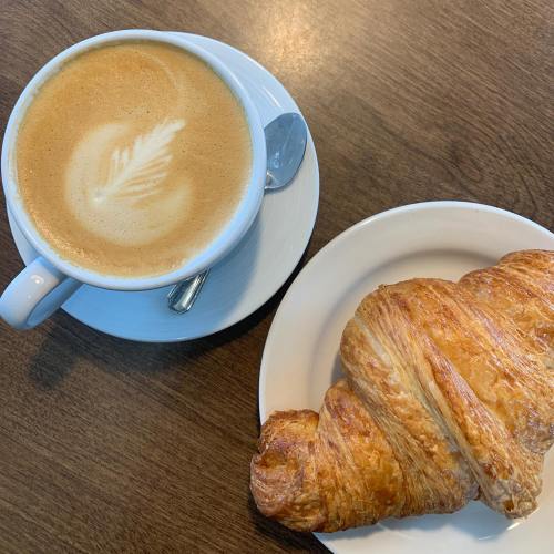 A cup of latte with a leaf design and a golden-brown croissant on a white plate, set on a wooden table.