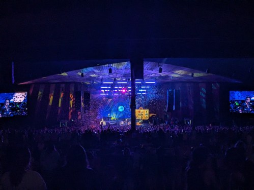 A lively concert scene with a large crowd, colorful lights, and confetti falling from the stage.