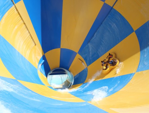 Two people ride a yellow and blue water slide, spiraling down into a circular tunnel. Water splashes around them.