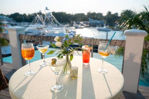 A table with five colorful cocktails and a small flower arrangement, overlooking a marina at sunset.