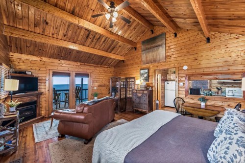 Cozy wooden cabin interior with a bed, sofa, and large windows showcasing a scenic view. Warm, rustic decor.