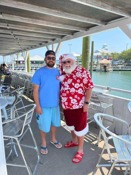 A man in a blue shirt and shorts stands next to a Santa in a floral shirt and red shorts by a marina.