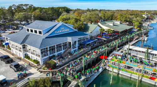 Aerial view of The Claw House restaurant with crowds, boats, and greenery along the waterfront on a sunny day.