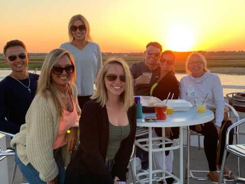 A group of seven people smiling and posing together on a boat during sunset, with drinks on a table.