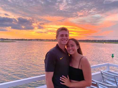 A couple smiles together on a boat at sunset, with vibrant orange and purple skies reflecting on the water.