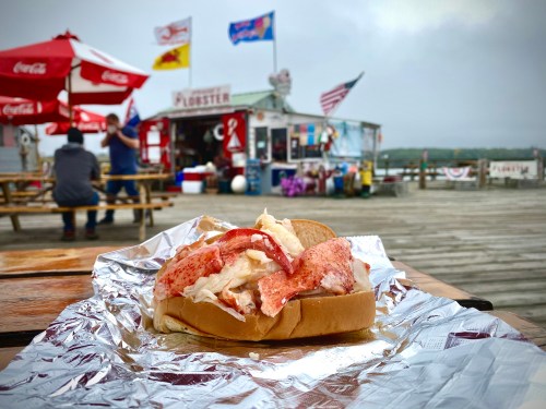 A lobster roll with lobster meat and coleslaw on a bun, set on a table near a seafood shack by the water.